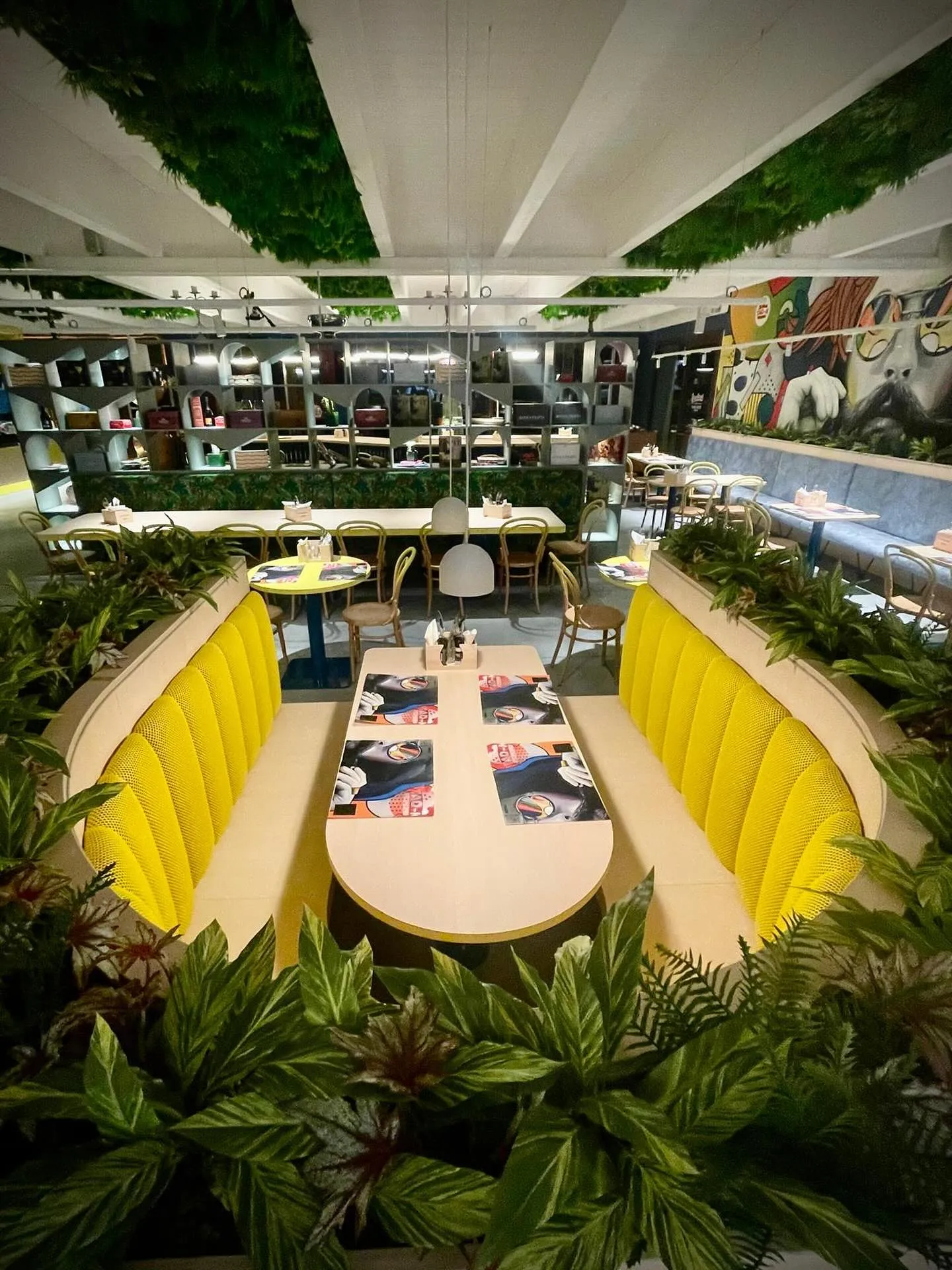 Interno del locale con tavoli, sedute gialle, molte piante decorative e illuminazione moderna che crea un ambiente accogliente