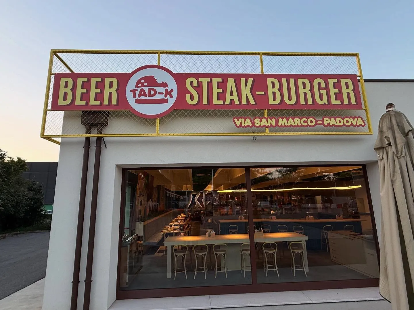 Facciata del ristorante con insegna TAD-K e scritta Beer Steak Burger, con tavoli e sala interna visibili attraverso la vetrina